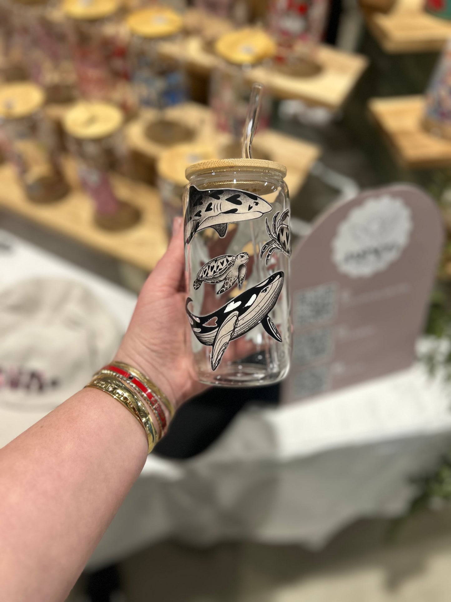 Hand holding a clear glass cup with whale design and wooden lid, blurred background