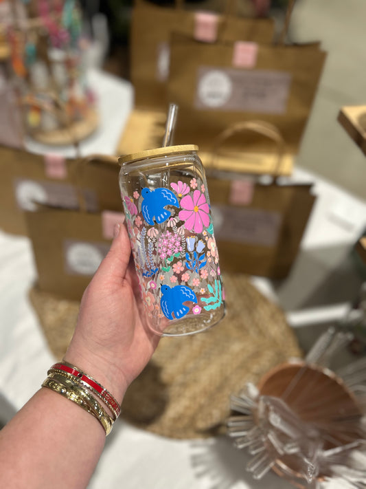Hand holding a decorated glass tumbler with floral and animal design, surrounded by brown boxes.