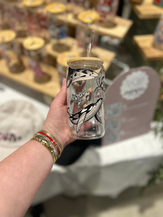 Hand holding a clear glass cup with whale design and wooden lid, blurred background