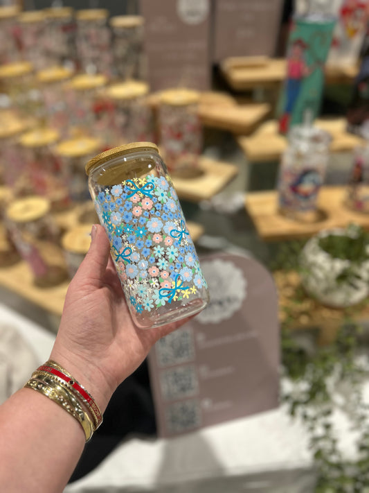 Hand holding a glass cup with blue decorative flowers and bows, bamboo lid, blurred background